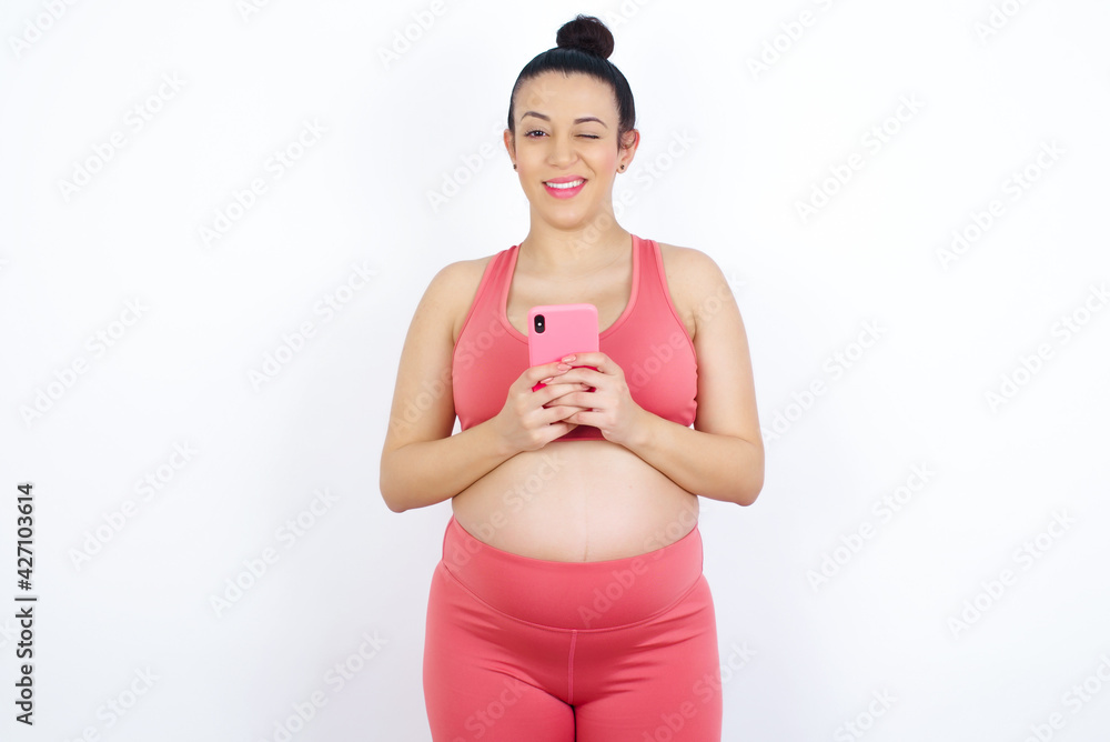 Pleased young beautiful Arab pregnant woman in sports clothes against white wall using self phone and looking and winking at the camera. Flirt and coquettish concept. Pleased young beautiful Arab pregnant woman in sports clothes against white wall using self phone and looking and winking at the camera. Flirt and coquettish concept.