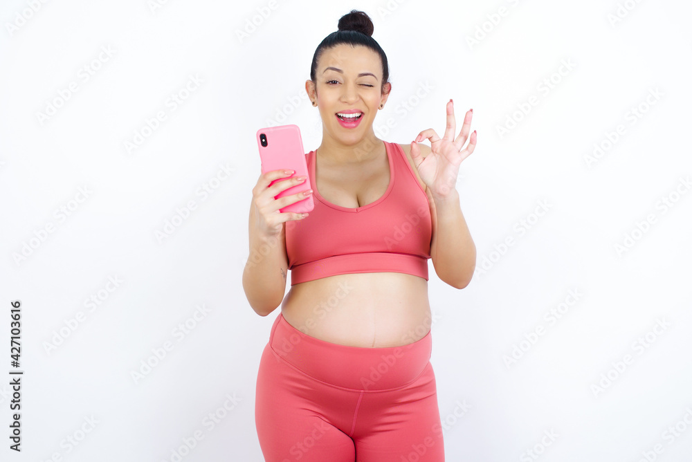 Happy young beautiful Arab pregnant woman in sports clothes against white wall sending a message on his smartphone or taking a selfie and making ok sign with his hand. Happy young beautiful Arab pregnant woman in sports clothes against white wall sending a message on his smartphone or taking a selfie and making ok sign with his hand.