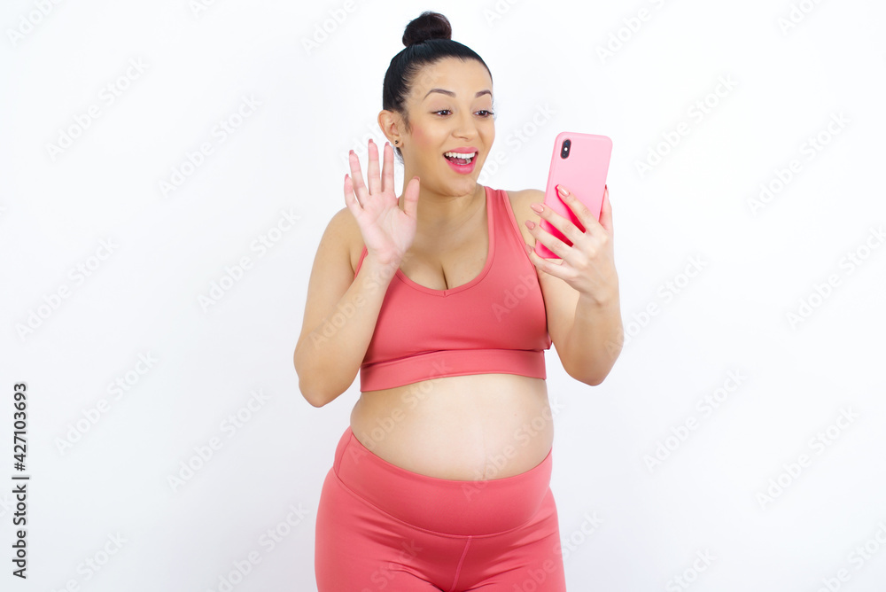 Portrait of happy friendly young beautiful Arab pregnant woman in sports clothes against white wall taking selfie and waving hand, communicating on video call, online chatting. Portrait of happy friendly young beautiful Arab pregnant woman in sports clothes against white wall taking selfie and waving hand, communicating on video call, online chatting.