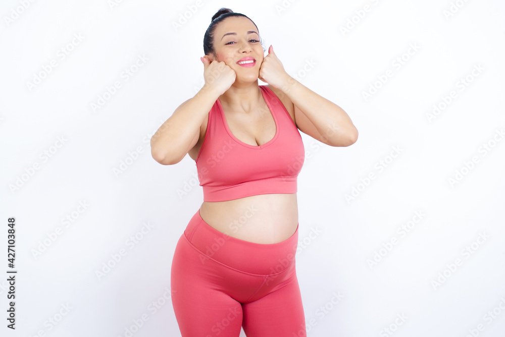 Happy young beautiful Arab pregnant woman in sports clothes against white wall keeps fists on cheeks smiles broadly and has positive expression being in good mood Happy young beautiful Arab pregnant woman in sports clothes against white wall keeps fists on cheeks smiles broadly and has positive expression being in good mood