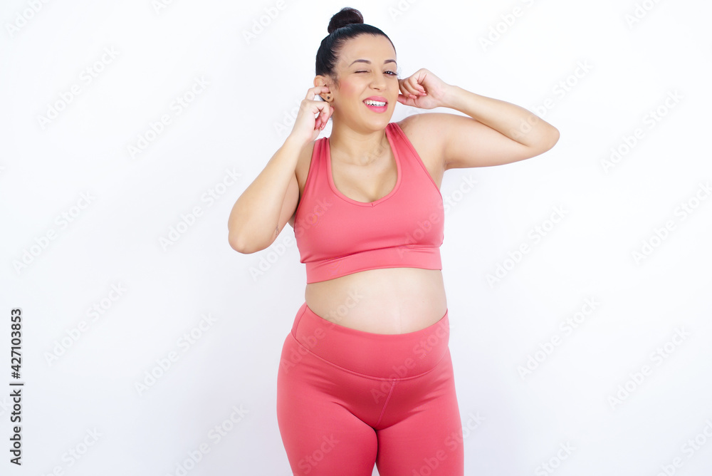 Happy young beautiful Arab pregnant woman in sports clothes against white wall ignores loud music and plugs ears with fingers asks to turn off sound Happy young beautiful Arab pregnant woman in sports clothes against white wall ignores loud music and plugs ears with fingers asks to turn off sound