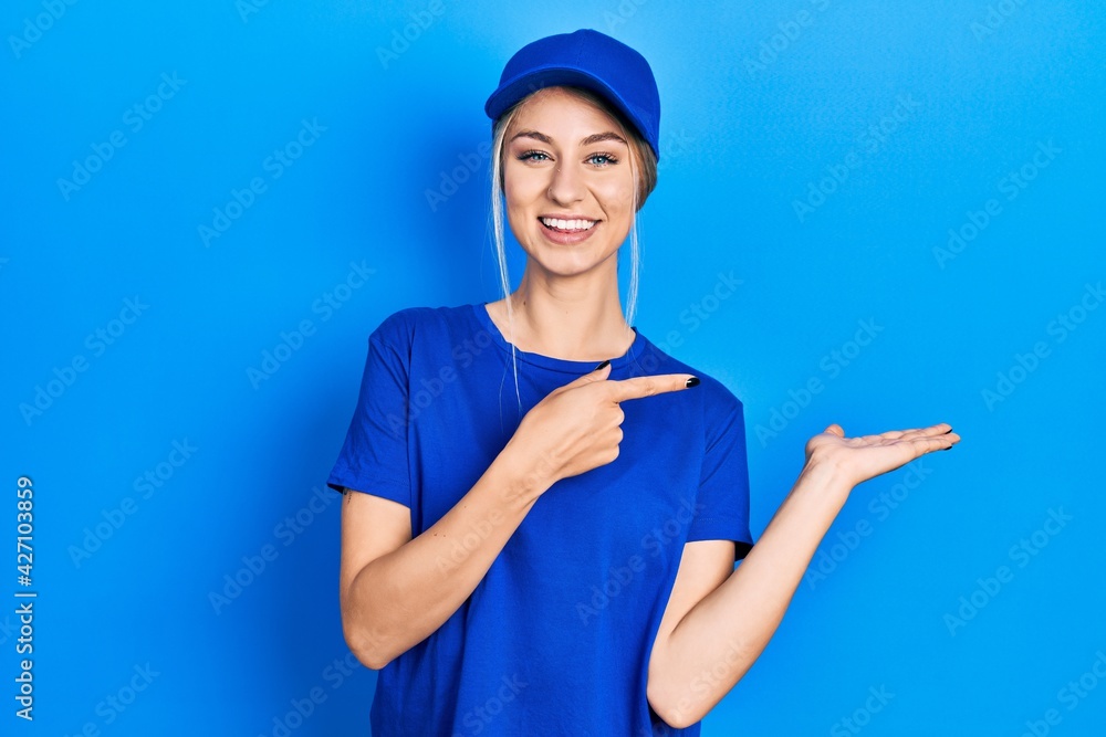 Young caucasian woman wearing courier uniform wearing cap amazed and smiling to the camera while presenting with hand and pointing with finger. Young caucasian woman wearing courier uniform wearing cap amazed and smiling to the camera while presenting with hand and pointing with finger.