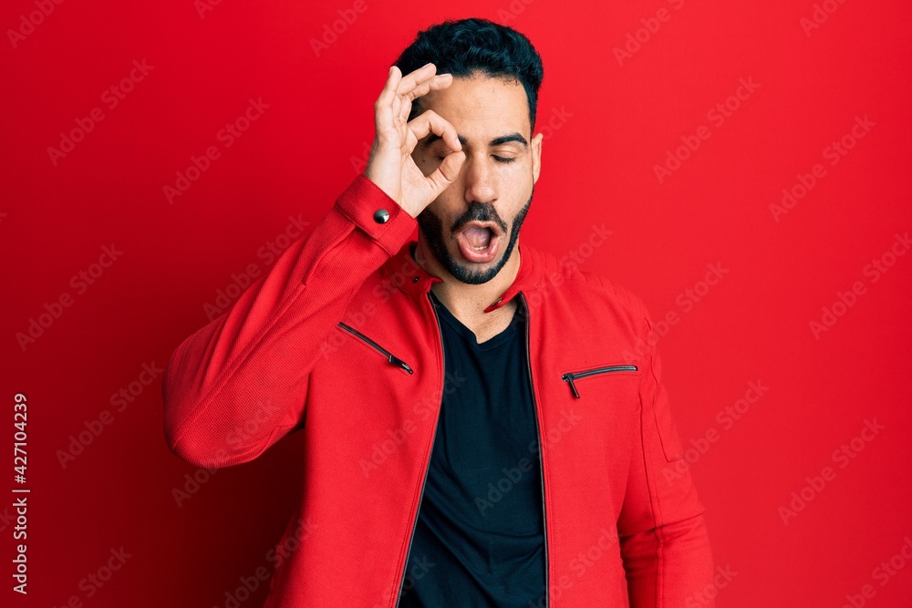 Young hispanic man wearing red leather jacket doing ok gesture shocked with surprised face, eye looking through fingers. unbelieving expression. Young hispanic man wearing red leather jacket doing ok gesture shocked with surprised face, eye looking through fingers. unbelieving expression.