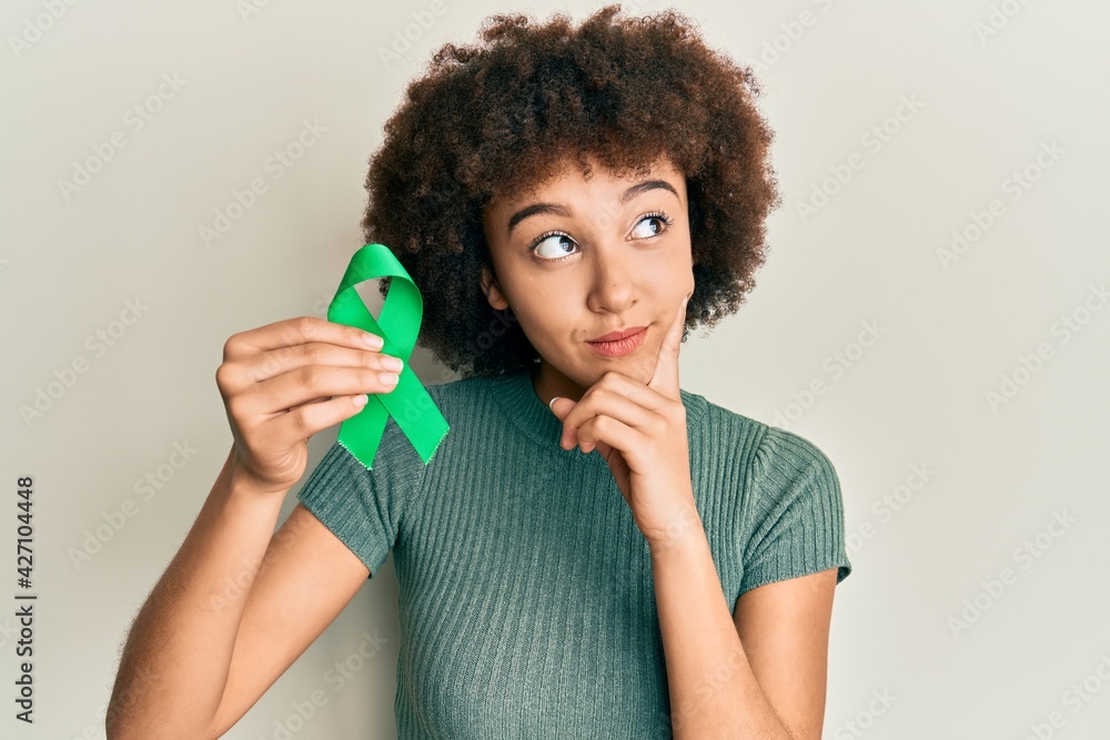 Young hispanic girl holding support green ribbon serious face thinking about question with hand on chin, thoughtful about confusing idea Young hispanic girl holding support green ribbon serious face thinking about question with hand on chin, thoughtful about confusing idea
