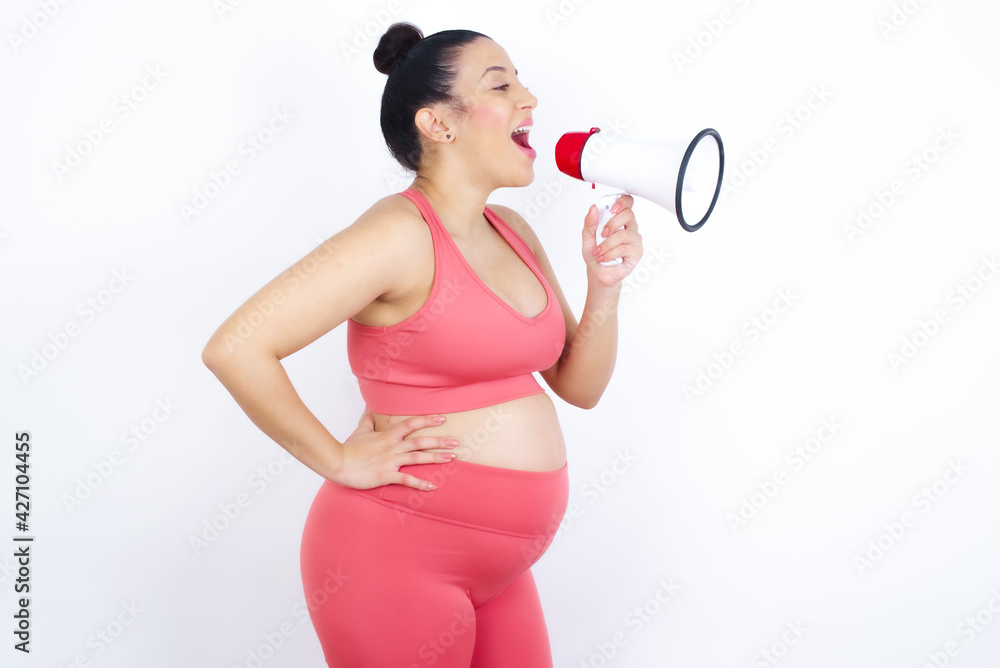 Funny young beautiful Arab pregnant woman in sports clothes against white wall People sincere emotions lifestyle concept. Mock up copy space. Screaming in megaphone. Funny young beautiful Arab pregnant woman in sports clothes against white wall People sincere emotions lifestyle concept. Mock up copy space. Screaming in megaphone.