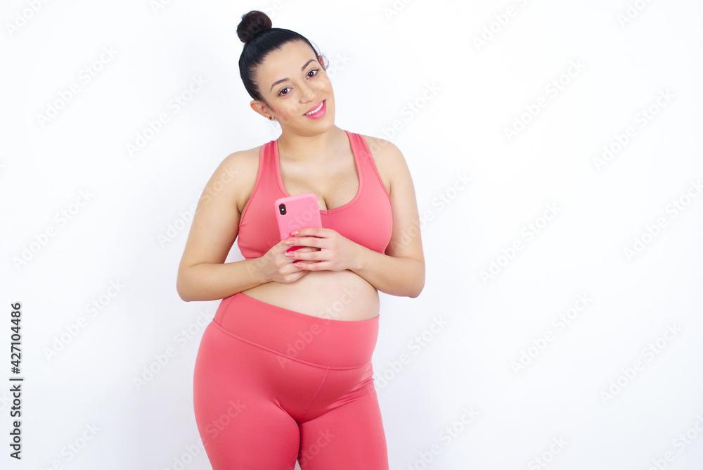 young beautiful Arab pregnant woman in sports clothes against white wall Mock up copy space. Using mobile phone, typing sms message young beautiful Arab pregnant woman in sports clothes against white wall Mock up copy space. Using mobile phone, typing sms message
