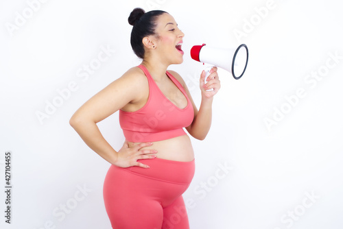Funny young beautiful Arab pregnant woman in sports clothes against white wall People sincere emotions lifestyle concept. Mock up copy space. Screaming in megaphone.