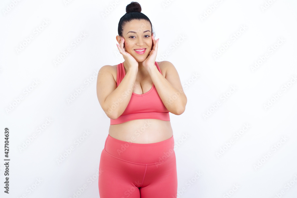 Happy young beautiful Arab pregnant woman in sports clothes against white wall touches both cheeks gently, has tender smile, shows white teeth, gazes positively straightly at camera, Happy young beautiful Arab pregnant woman in sports clothes against white wall touches both cheeks gently, has tender smile, shows white teeth, gazes positively straightly at camera,