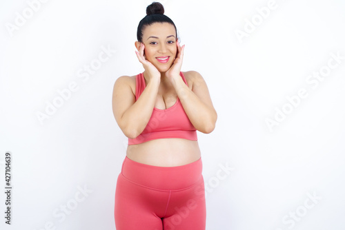 Fototapeta Naklejka Na Ścianę i Meble -  Happy young beautiful Arab pregnant woman in sports clothes against white wall touches both cheeks gently, has tender smile, shows white teeth, gazes positively straightly at camera,