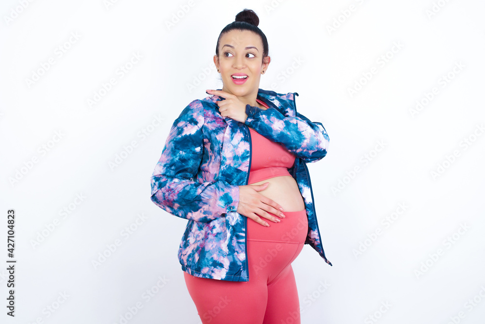 Cheerful young beautiful Arab pregnant woman in sports clothes against white wall demonstrating hairdo Cheerful young beautiful Arab pregnant woman in sports clothes against white wall demonstrating hairdo