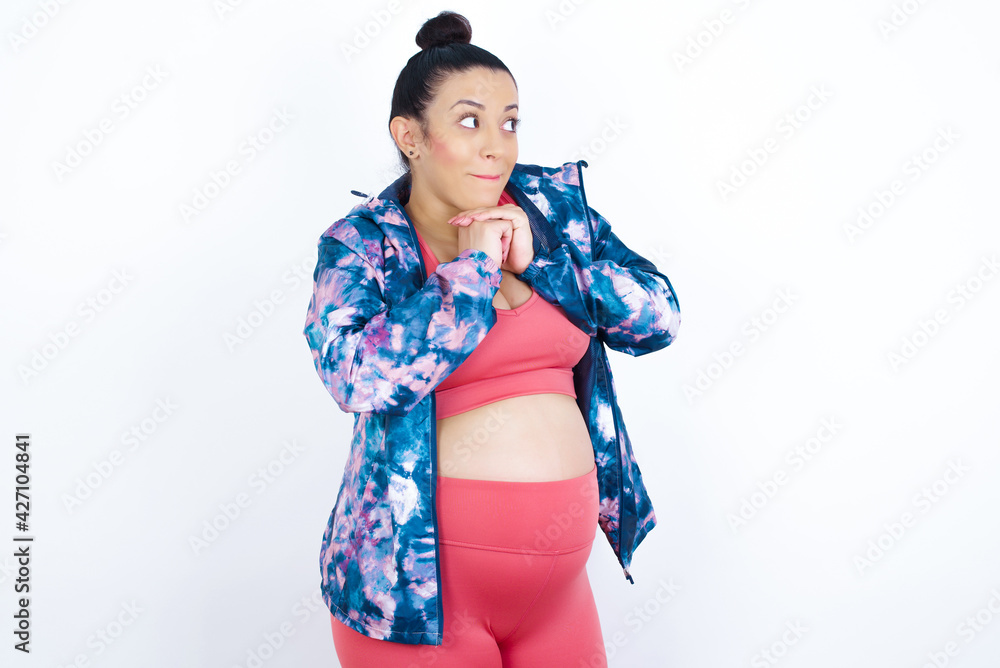 Curious young beautiful Arab pregnant woman in sports clothes against white wall keeps hands under chin bites lips and looks with interest aside. Curious young beautiful Arab pregnant woman in sports clothes against white wall keeps hands under chin bites lips and looks with interest aside.