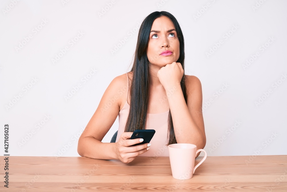 Young caucasian woman sitting at the desk using smartphone drinking coffee serious face thinking about question with hand on chin, thoughtful about confusing idea Young caucasian woman sitting at the desk using smartphone drinking coffee serious face thinking about question with hand on chin, thoughtful about confusing idea