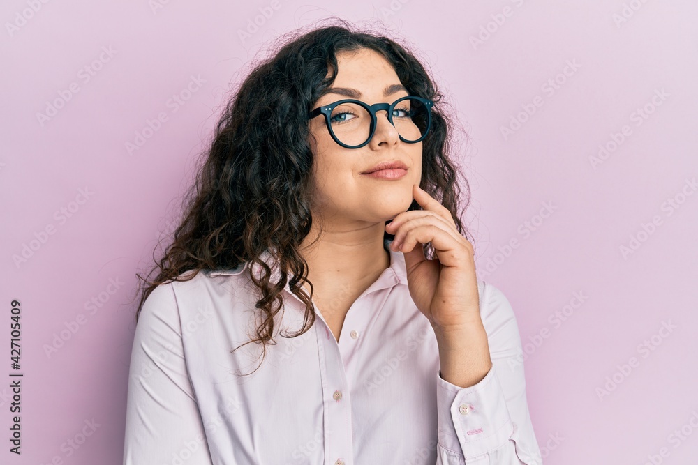 Young brunette woman with curly hair wearing casual clothes and glasses looking confident at the camera with smile with crossed arms and hand raised on chin. thinking positive.