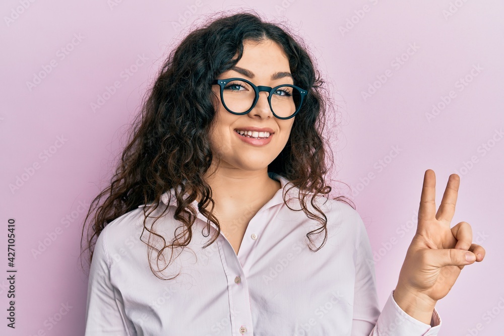 Young brunette woman with curly hair wearing casual clothes and glasses showing and pointing up with fingers number two while smiling confident and happy. Young brunette woman with curly hair wearing casual clothes and glasses showing and pointing up with fingers number two while smiling confident and happy.