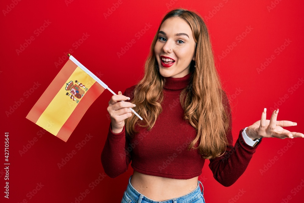 Young blonde woman holding spain flag celebrating achievement with happy smile and winner expression with raised hand Young blonde woman holding spain flag celebrating achievement with happy smile and winner expression with raised hand