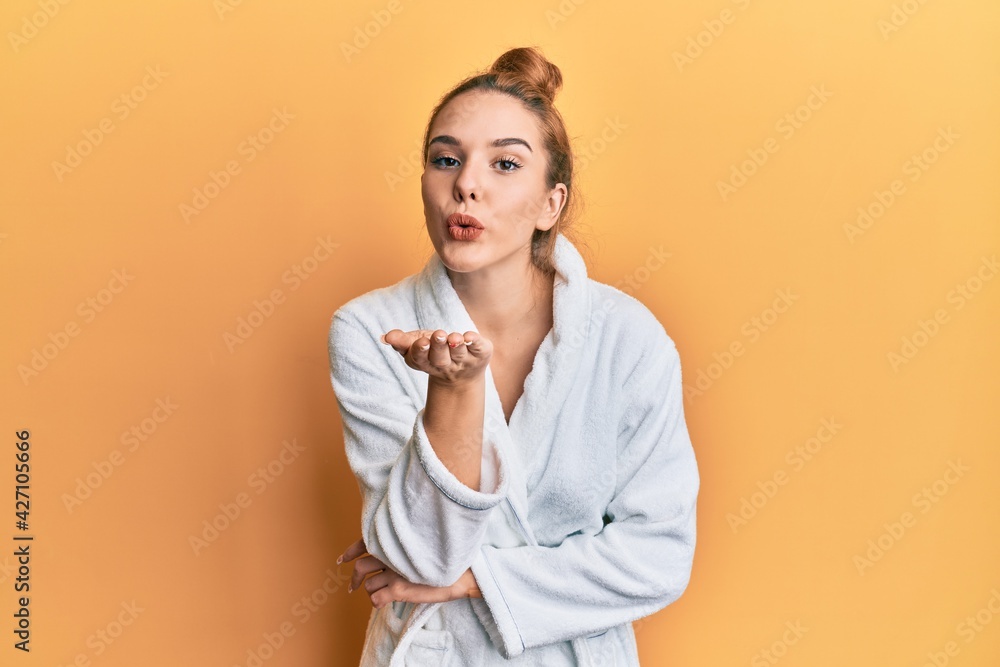 Young blonde woman wearing bathrobe looking at the camera blowing a kiss with hand on air being lovely and sexy. love expression. Young blonde woman wearing bathrobe looking at the camera blowing a kiss with hand on air being lovely and sexy. love expression.