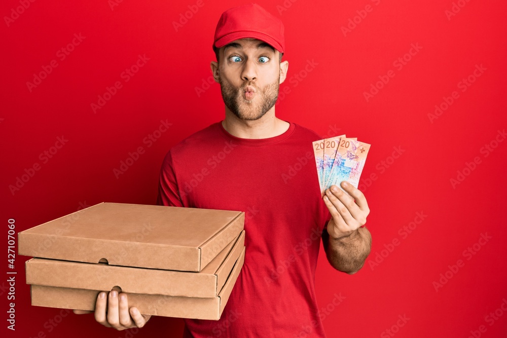 Young caucasian man holding delivery box and swiss franc banknotes making fish face with mouth and squinting eyes, crazy and comical. Young caucasian man holding delivery box and swiss franc banknotes making fish face with mouth and squinting eyes, crazy and comical.