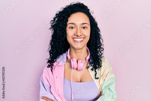 Young hispanic woman with curly hair wearing gym clothes and using headphones happy face smiling with crossed arms looking at the camera. positive person.