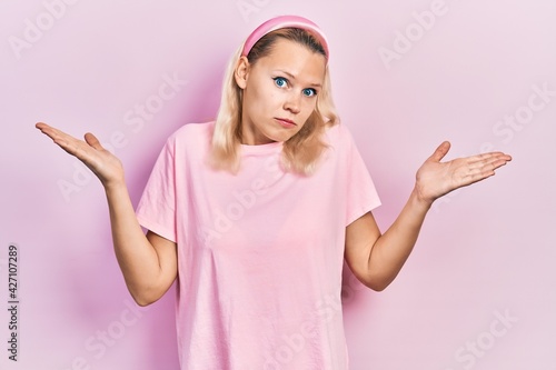 Beautiful caucasian blonde woman wearing casual pink t shirt clueless and confused with open arms, no idea and doubtful face. photo