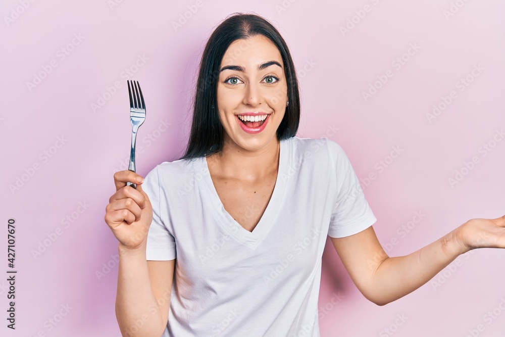 Beautiful woman with blue eyes holding one silver fork celebrating achievement with happy smile and winner expression with raised hand Beautiful woman with blue eyes holding one silver fork celebrating achievement with happy smile and winner expression with raised hand