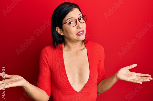 Young latin woman wearing casual clothes and glasses clueless and confused expression with arms and hands raised. doubt concept.