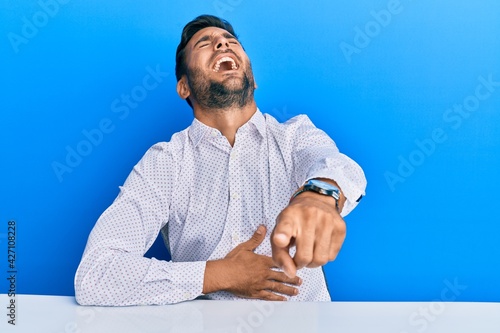 Handsome hispanic man wearing business clothes sitting on the table laughing at you, pointing finger to the camera with hand over body, shame expression photo