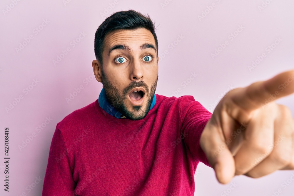 Young hispanic man wearing casual clothes pointing with finger surprised ahead, open mouth amazed expression, something on the front Young hispanic man wearing casual clothes pointing with finger surprised ahead, open mouth amazed expression, something on the front