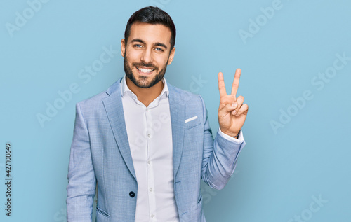 Young hispanic man wearing business jacket smiling with happy face winking at the camera doing victory sign. number two.