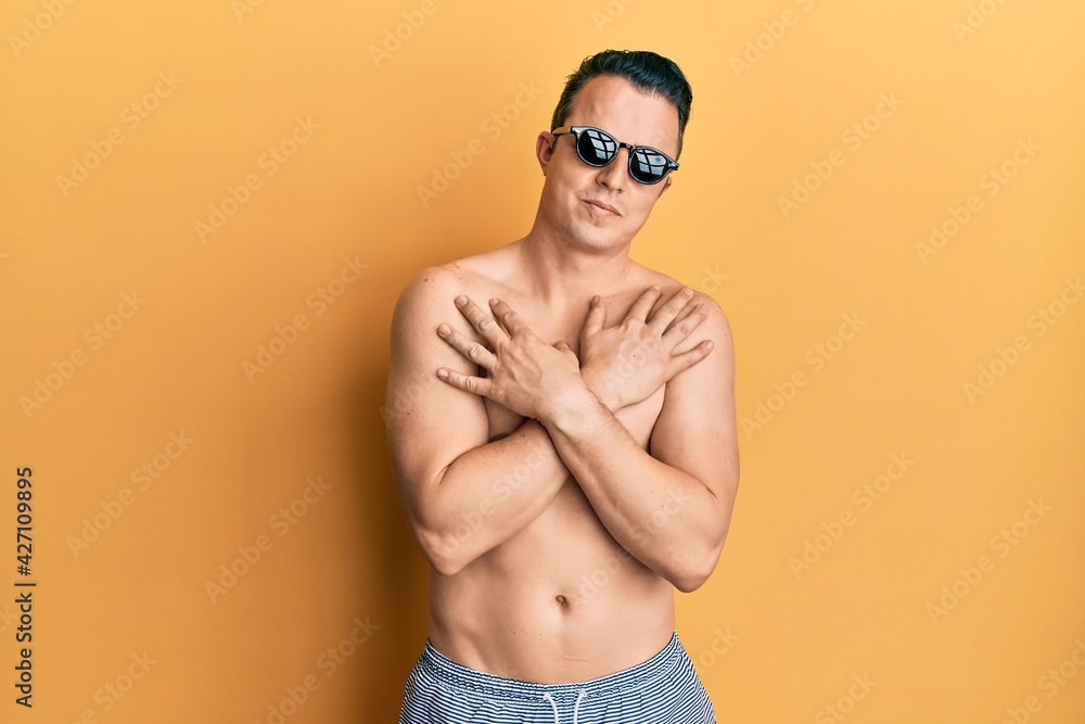 Fototapeta premium Handsome young man wearing swimsuit and sunglasses hugging oneself happy and positive, smiling confident. self love and self care
