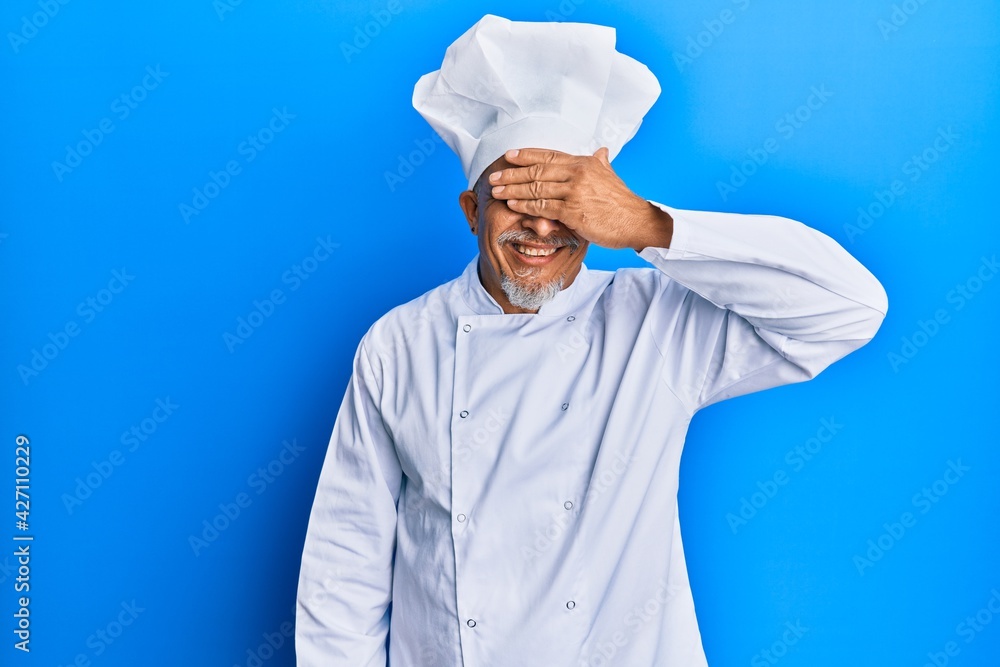 Middle age grey-haired man wearing professional cook uniform and hat smiling and laughing with hand on face covering eyes for surprise. blind concept. Middle age grey-haired man wearing professional cook uniform and hat smiling and laughing with hand on face covering eyes for surprise. blind concept.