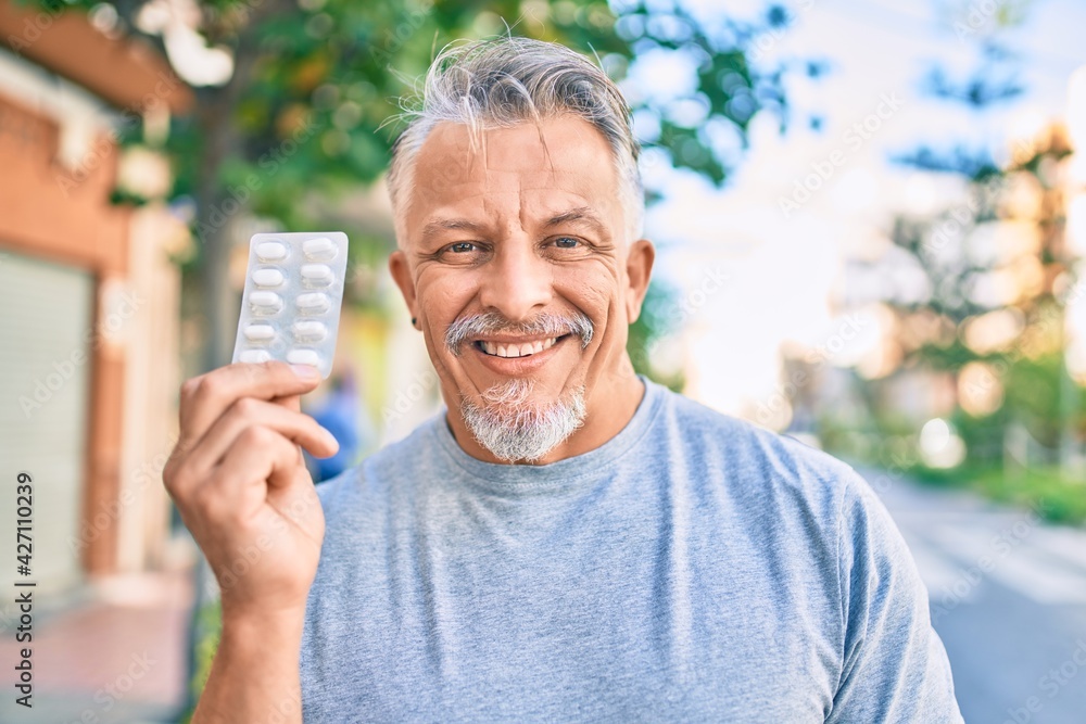 Middle age hispanic grey-haired man smiling happy holding pills standing at the city. Middle age hispanic grey-haired man smiling happy holding pills standing at the city.