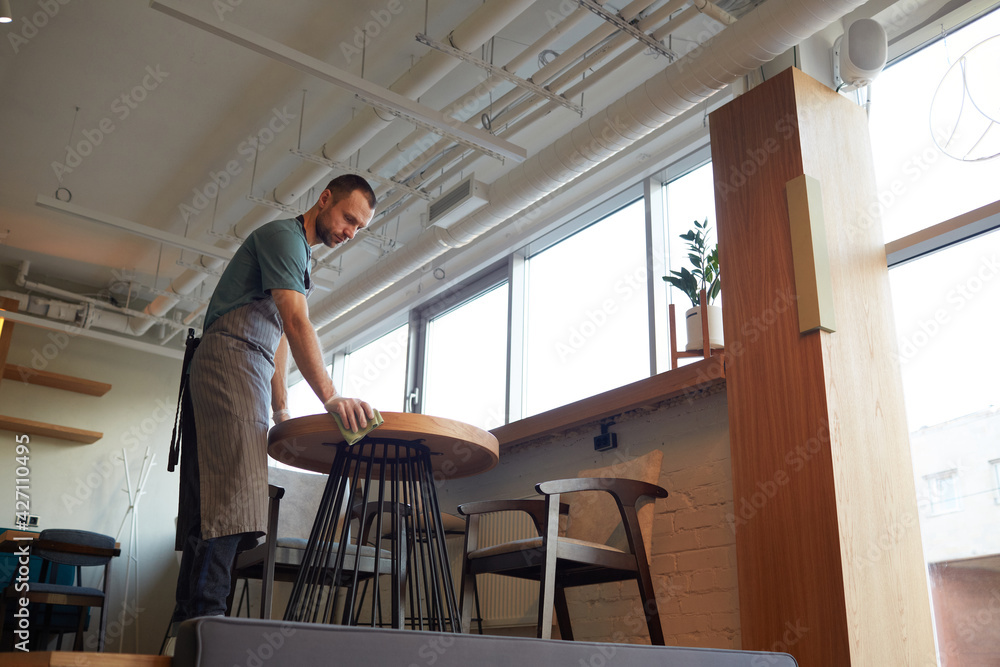 Low angle portrait of male waiter cleaning tables in cafe or coffee shop while preparing for opening in morning, copy space