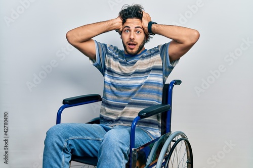 Handsome hispanic man sitting on wheelchair crazy and scared with hands on head, afraid and surprised of shock with open mouth