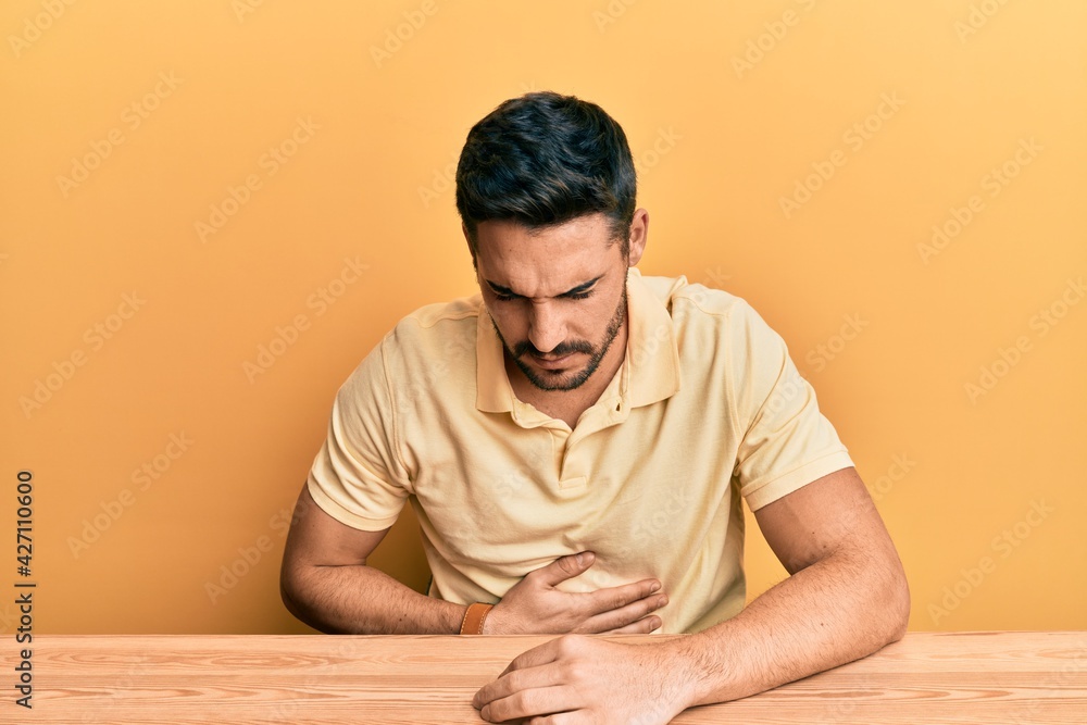 Young hispanic man wearing casual clothes sitting on the table with hand on stomach because nausea, painful disease feeling unwell. ache concept. Young hispanic man wearing casual clothes sitting on the table with hand on stomach because nausea, painful disease feeling unwell. ache concept.