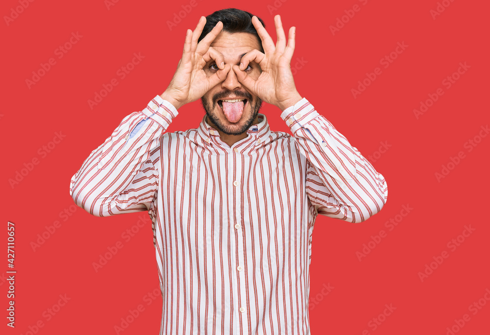 Young hispanic man wearing business shirt doing ok gesture like binoculars sticking tongue out, eyes looking through fingers. crazy expression. Young hispanic man wearing business shirt doing ok gesture like binoculars sticking tongue out, eyes looking through fingers. crazy expression.