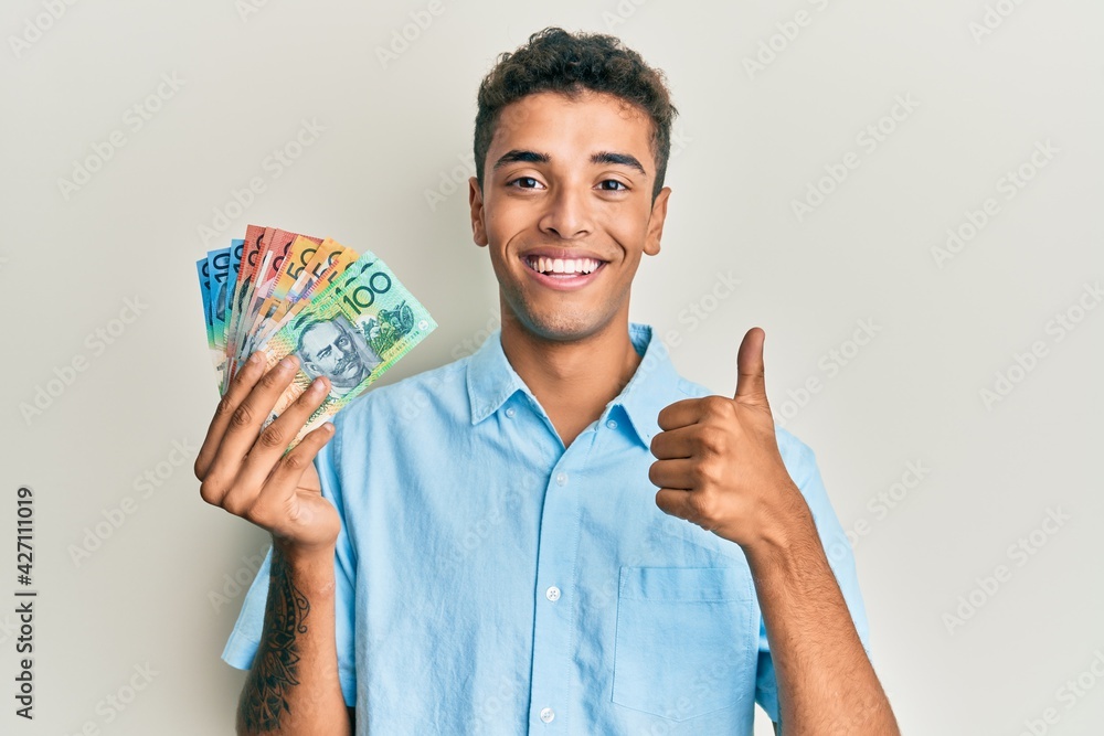 Young handsome african american man holding australian dollars smiling happy and positive, thumb up doing excellent and approval sign Young handsome african american man holding australian dollars smiling happy and positive, thumb up doing excellent and approval sign