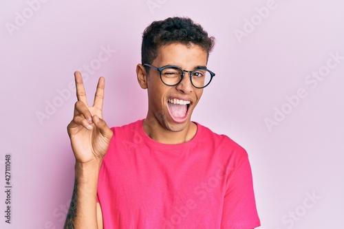 Young handsome african american man wearing glasses over pink background smiling with happy face winking at the camera doing victory sign. number two. © Krakenimages.com
