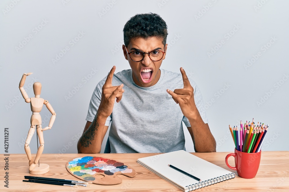 Naklejka premium Young handsome african american man painter sitting palette and art manikin shouting with crazy expression doing rock symbol with hands up. music star. heavy concept.