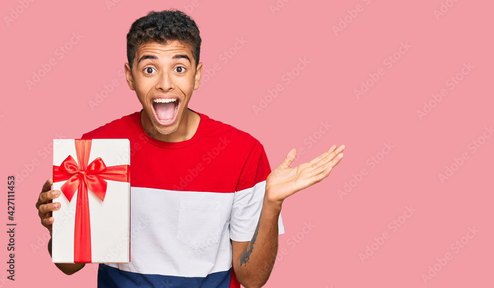 Young handsome african american man holding gift celebrating victory with happy smile and winner expression with raised hands Young handsome african american man holding gift celebrating victory with happy smile and winner expression with raised hands