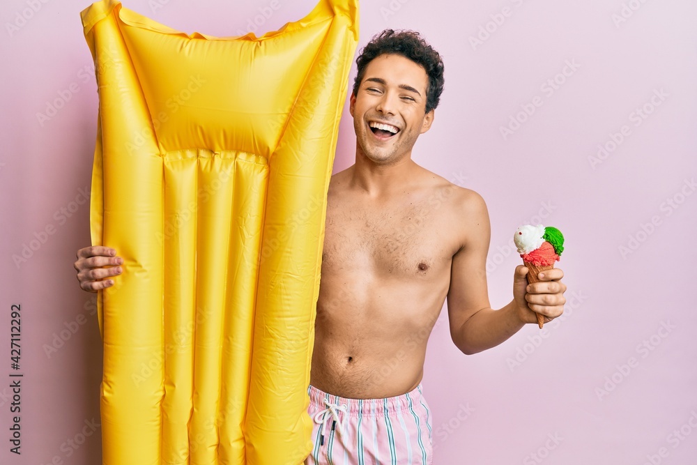 Obraz premium Young handsome man wearing swimsuit holding summer mattress float and icecream smiling and laughing hard out loud because funny crazy joke.