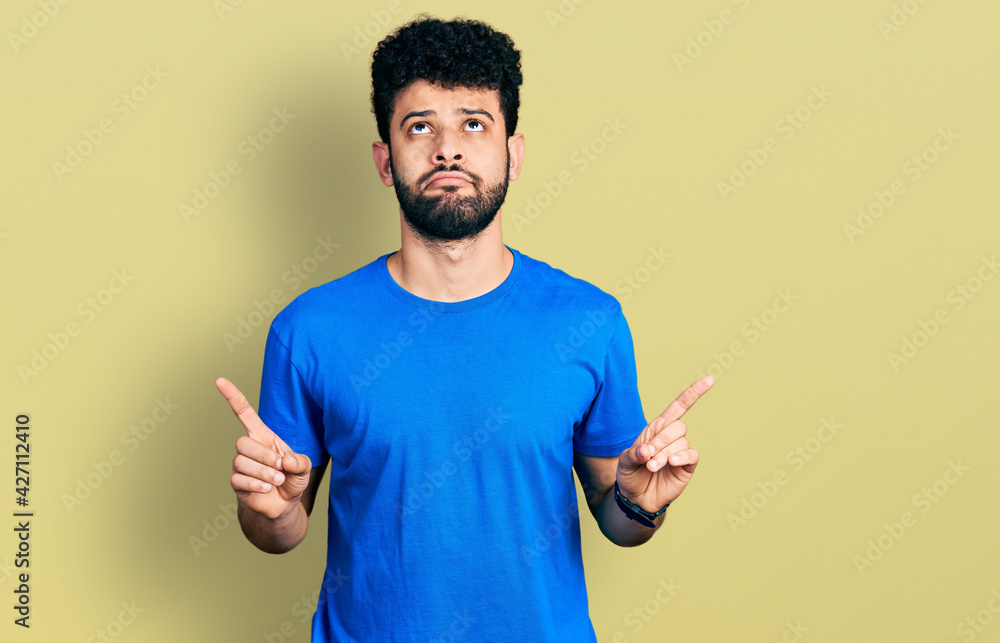 Young arab man with beard wearing casual blue t shirt pointing up looking sad and upset, indicating direction with fingers, unhappy and depressed. Young arab man with beard wearing casual blue t shirt pointing up looking sad and upset, indicating direction with fingers, unhappy and depressed.
