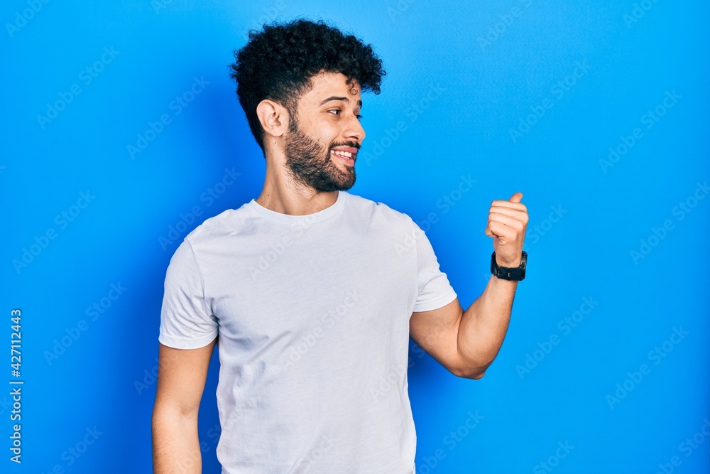 Young arab man with beard wearing casual white t shirt smiling with happy face looking and pointing to the side with thumb up. Young arab man with beard wearing casual white t shirt smiling with happy face looking and pointing to the side with thumb up.