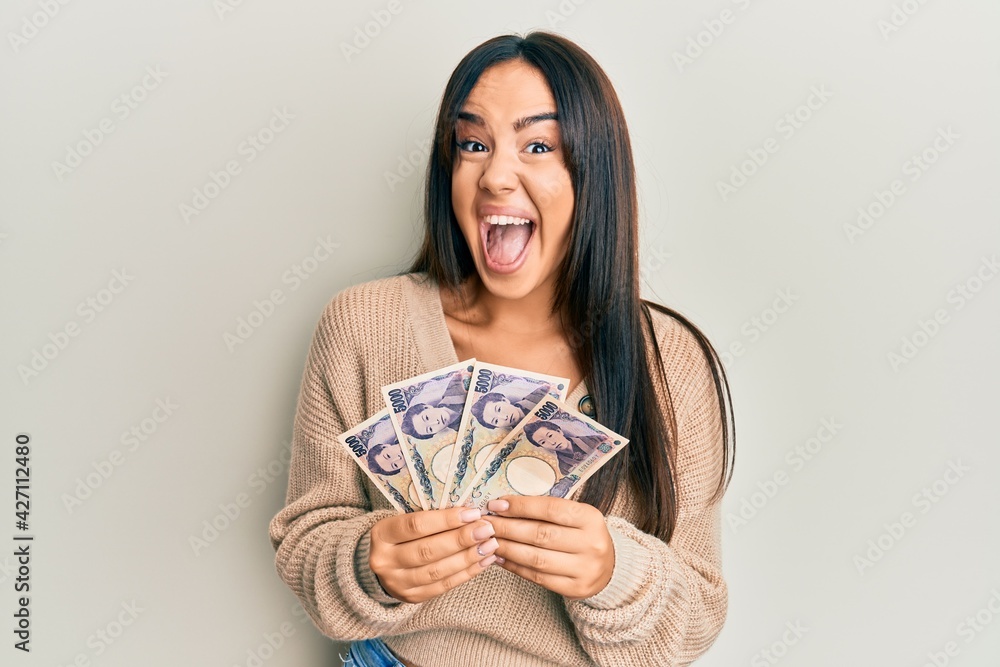 Young beautiful hispanic girl holding japanese yen banknotes celebrating crazy and amazed for success with open eyes screaming excited. Young beautiful hispanic girl holding japanese yen banknotes celebrating crazy and amazed for success with open eyes screaming excited.
