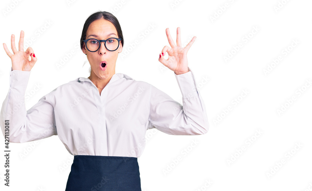 Beautiful brunette young woman wearing professional waitress apron looking surprised and shocked doing ok approval symbol with fingers. crazy expression Beautiful brunette young woman wearing professional waitress apron looking surprised and shocked doing ok approval symbol with fingers. crazy expression