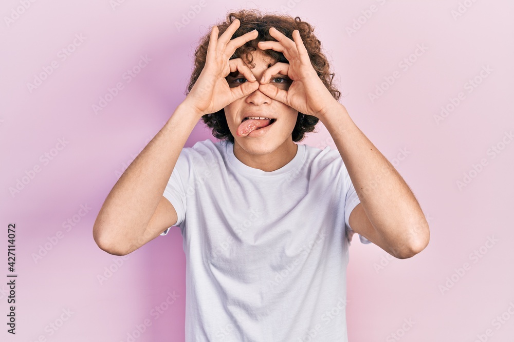 Handsome young man wearing casual white t shirt doing ok gesture like binoculars sticking tongue out, eyes looking through fingers. crazy expression. Handsome young man wearing casual white t shirt doing ok gesture like binoculars sticking tongue out, eyes looking through fingers. crazy expression.
