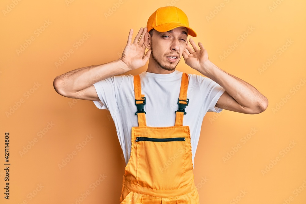 Hispanic young man wearing handyman uniform trying to hear both hands on ear gesture, curious for gossip. hearing problem, deaf Hispanic young man wearing handyman uniform trying to hear both hands on ear gesture, curious for gossip. hearing problem, deaf