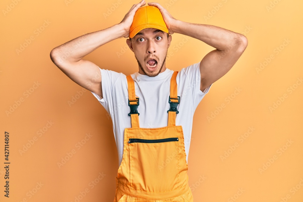 Hispanic young man wearing handyman uniform crazy and scared with hands on head, afraid and surprised of shock with open mouth