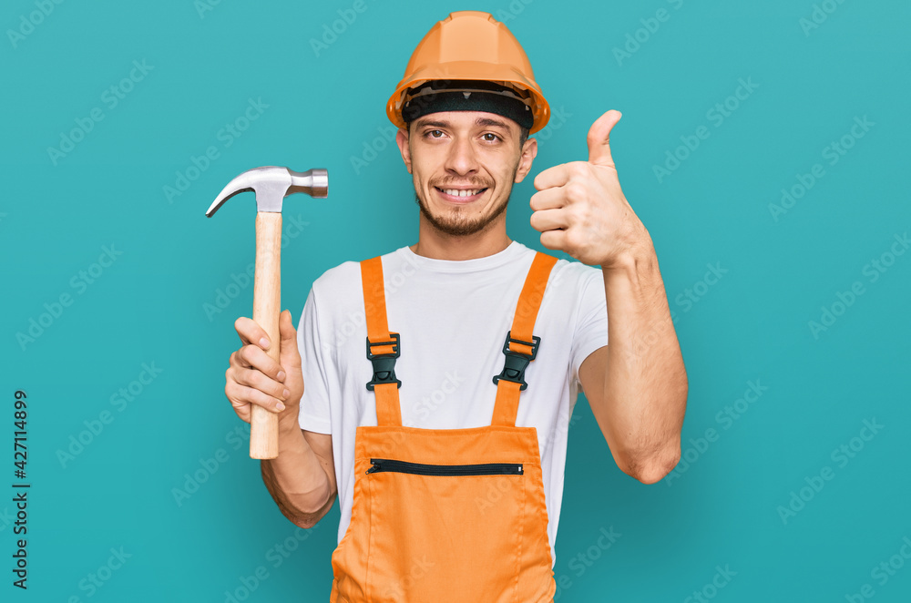 Hispanic young man wearing hardhat holding hammer smiling happy and positive, thumb up doing excellent and approval sign