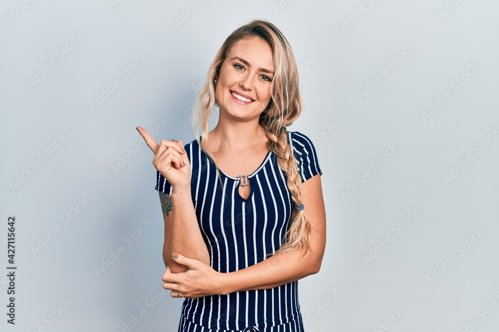 Beautiful young blonde woman wearing casual striped dress with a big smile on face, pointing with hand and finger to the side looking at the camera. Beautiful young blonde woman wearing casual striped dress with a big smile on face, pointing with hand and finger to the side looking at the camera.