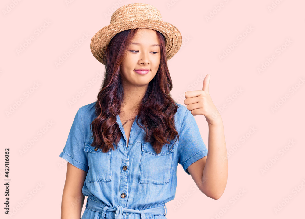 Young beautiful chinese girl wearing summer hat looking proud, smiling doing thumbs up gesture to the side Young beautiful chinese girl wearing summer hat looking proud, smiling doing thumbs up gesture to the side
